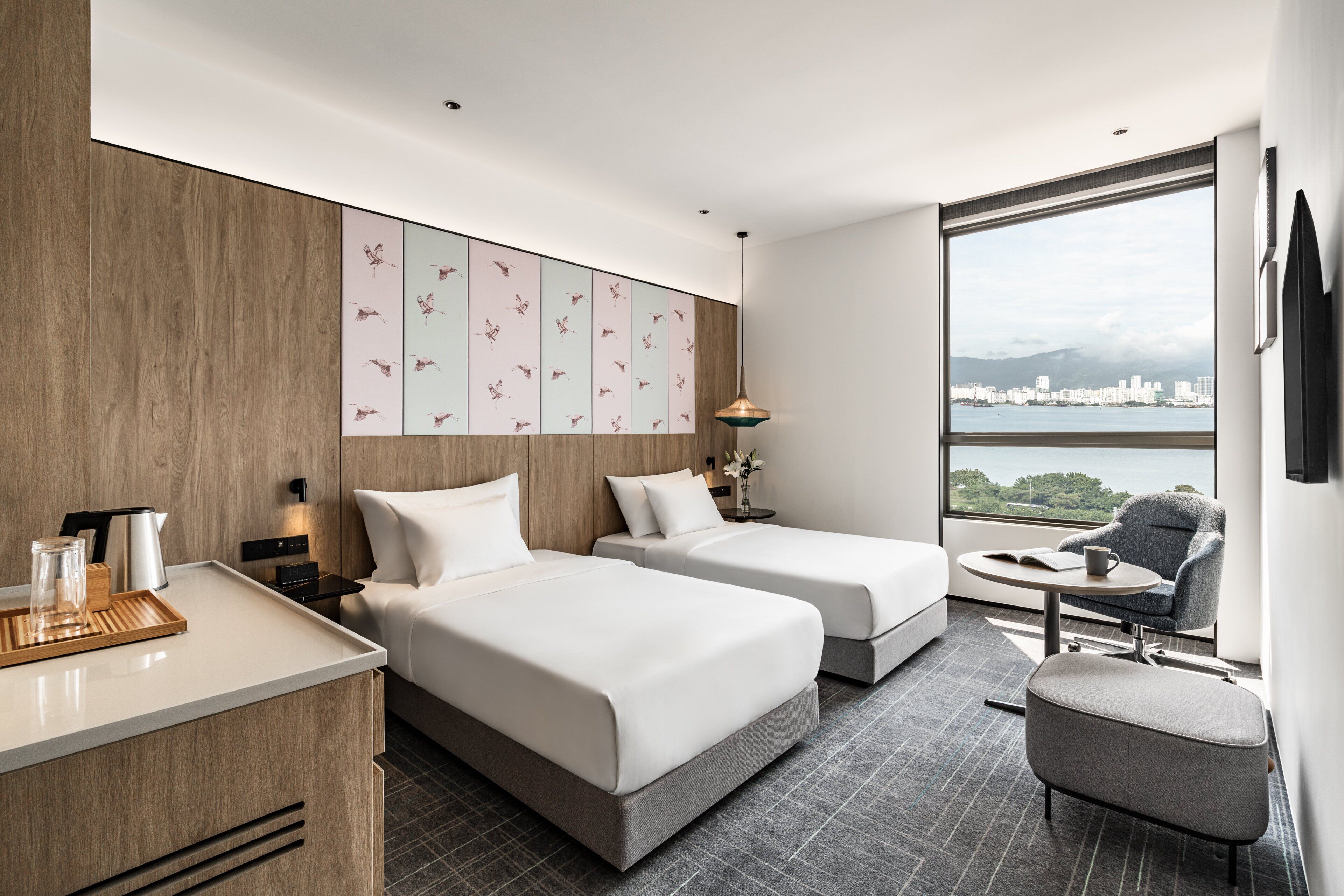 Ett modernt hotellrum med två sängar, ett skrivbord, en stol och ett stort fönster med utsikt över staden.