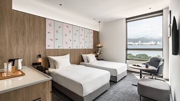 Ett modernt hotellrum med två sängar, ett skrivbord, en stol och ett stort fönster med utsikt över staden.