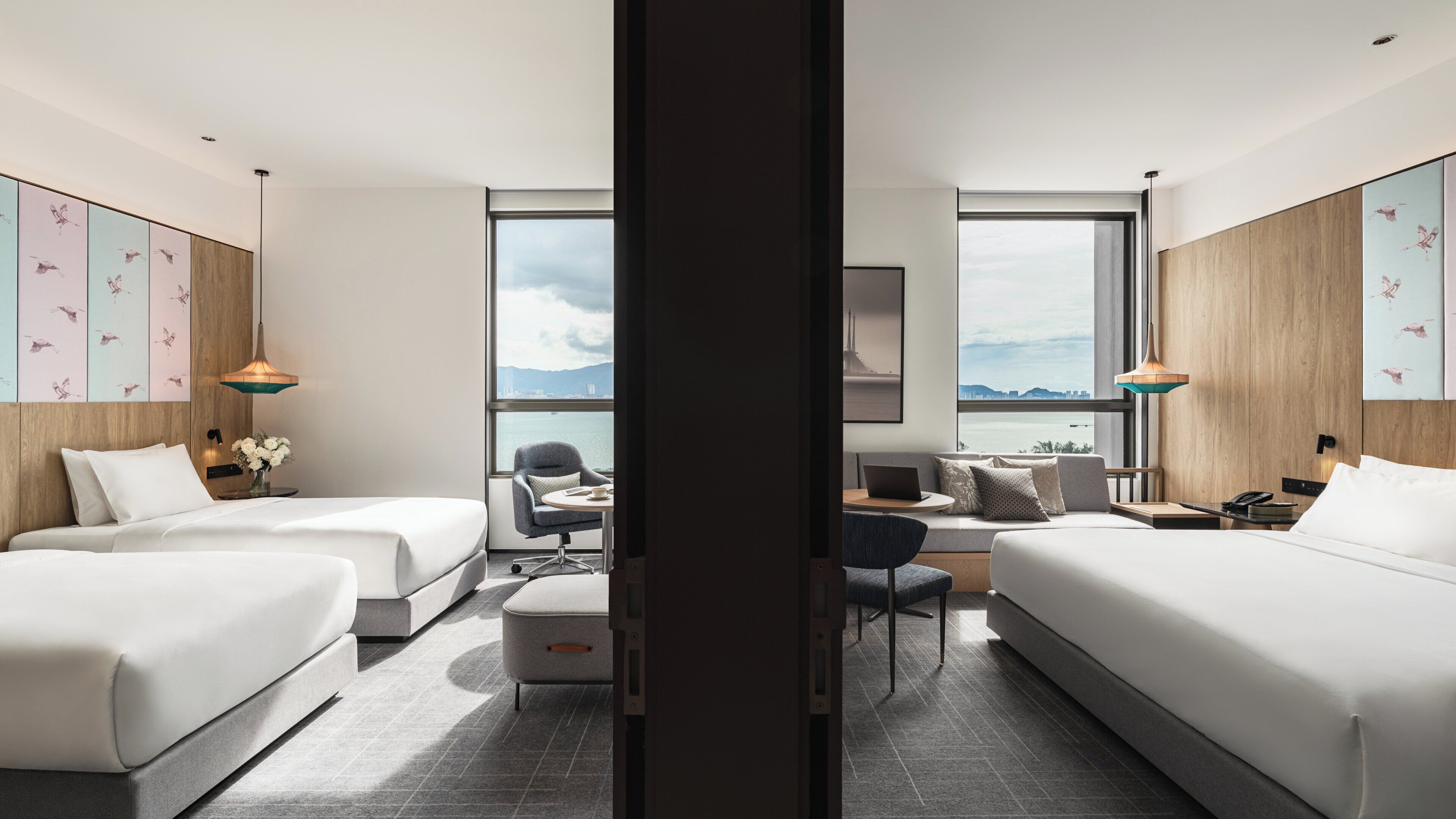 A modern hotel room with two beds, a desk, and a chair. There are large windows offering a view of the city.