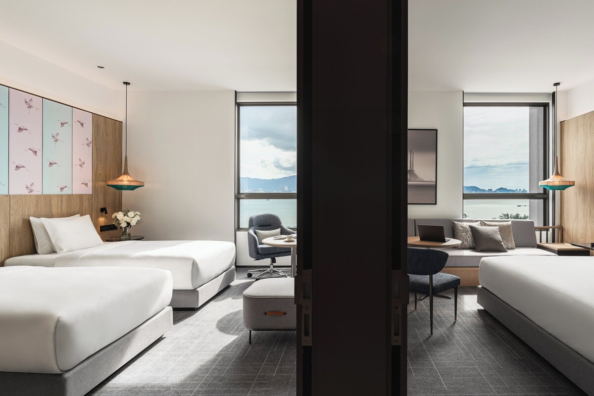 Habitación de hotel moderna con dos camas, un escritorio y una silla. Amplios ventanales ofrecen vistas a la ciudad.