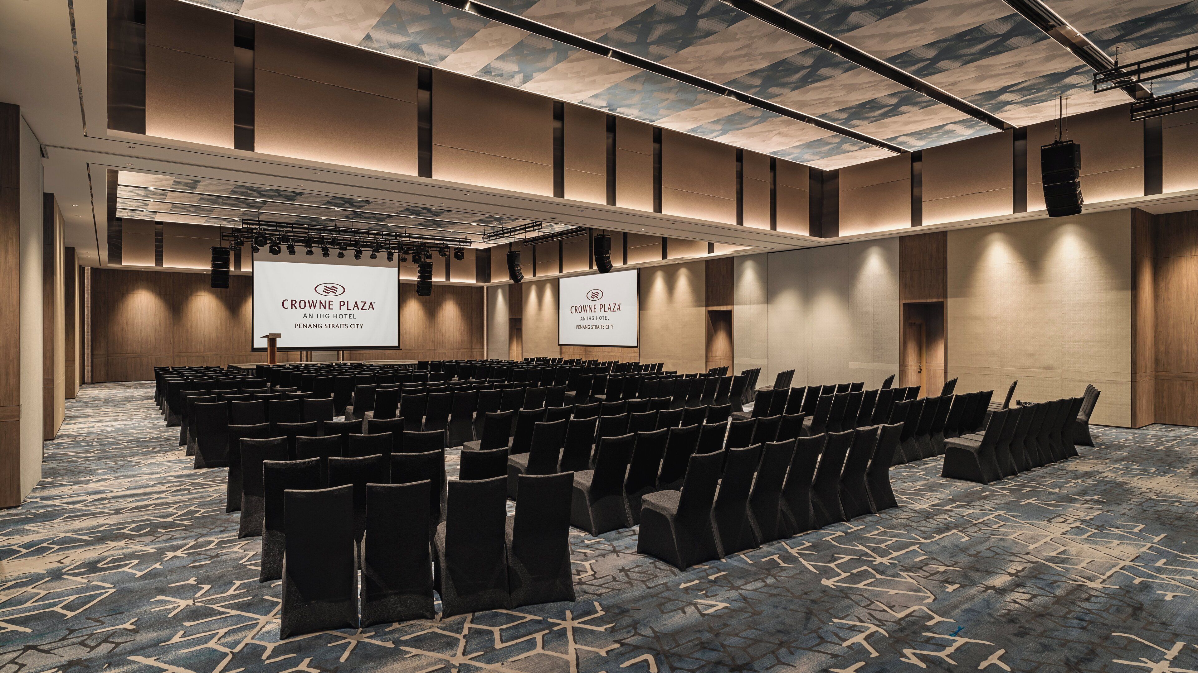 Un salón de conferencias en Crowne Plaza con filas de sillas, una pantalla grande y una alfombra estampada.