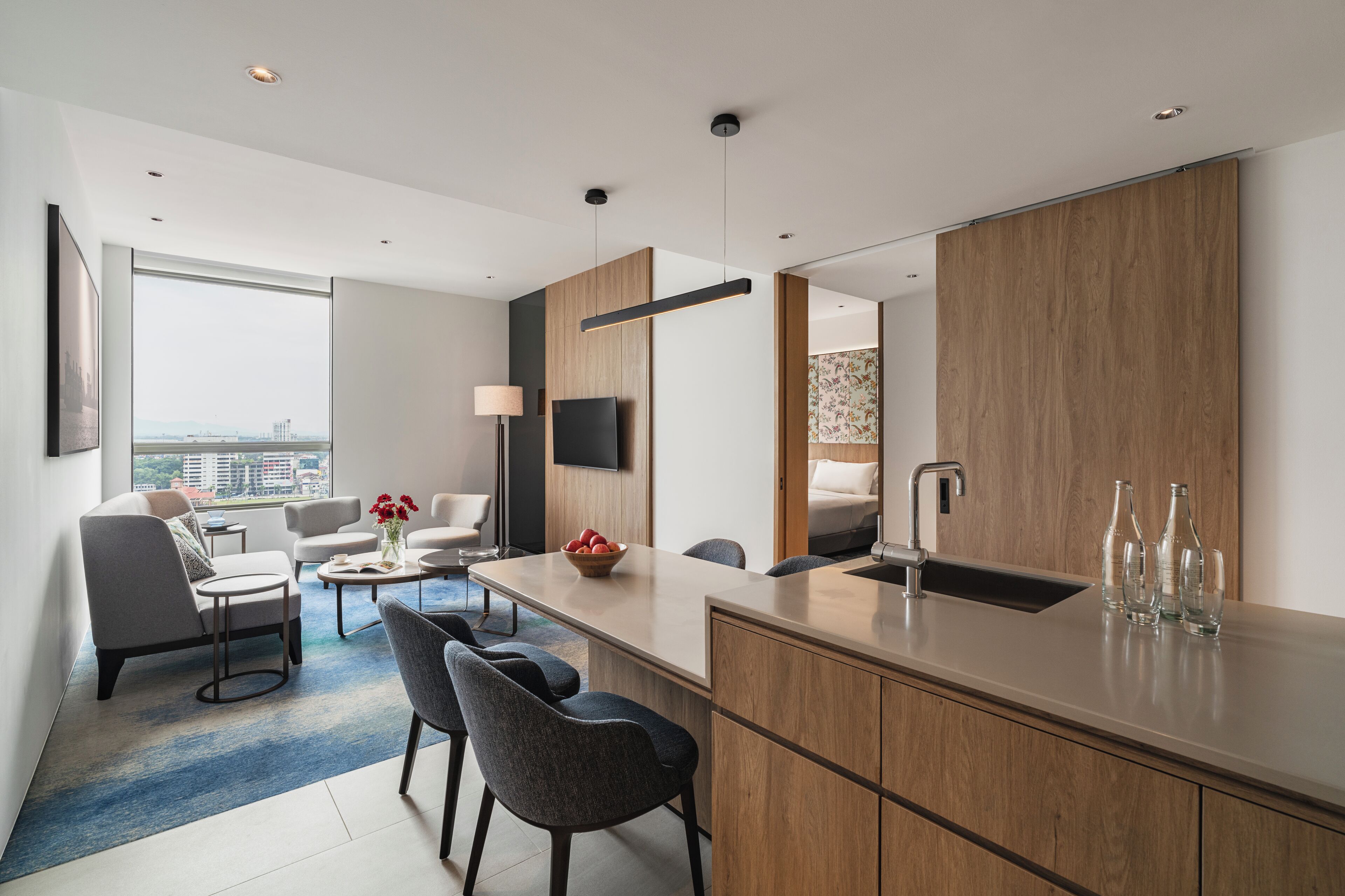 A modern hotel room with a large window, a flat-screen TV, a dining area, and a kitchen counter.