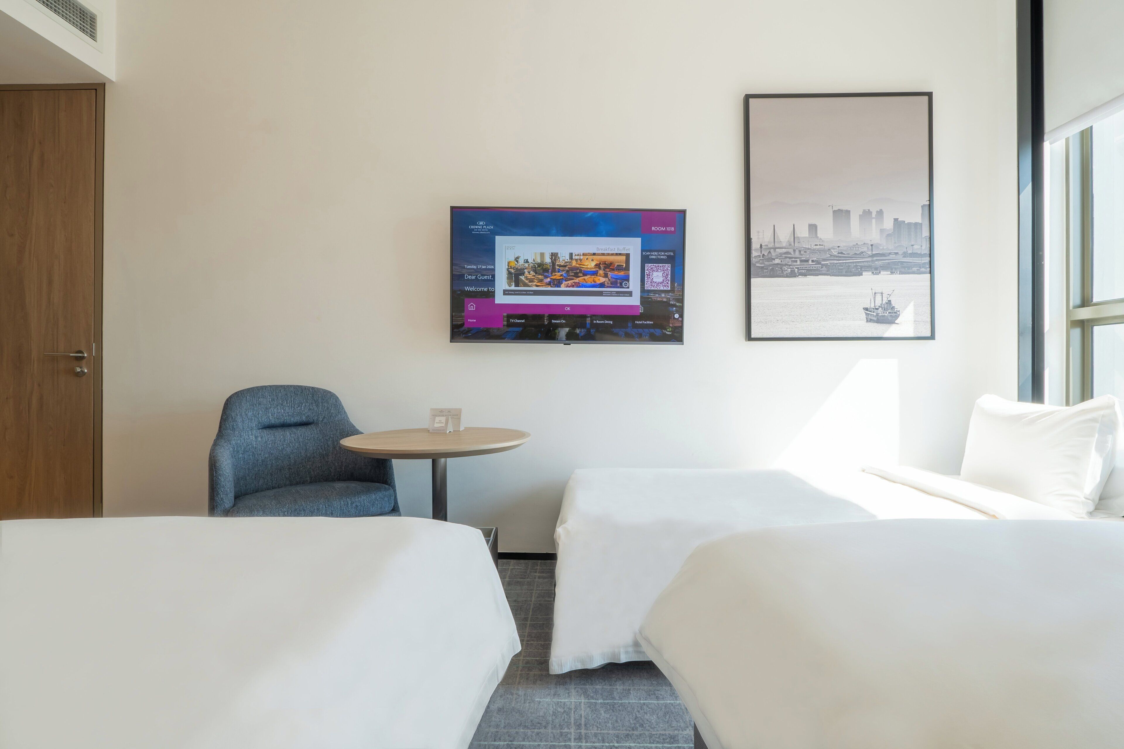 A hotel room with two beds, a small round table, a chair, a TV on the wall, and a framed cityscape picture.