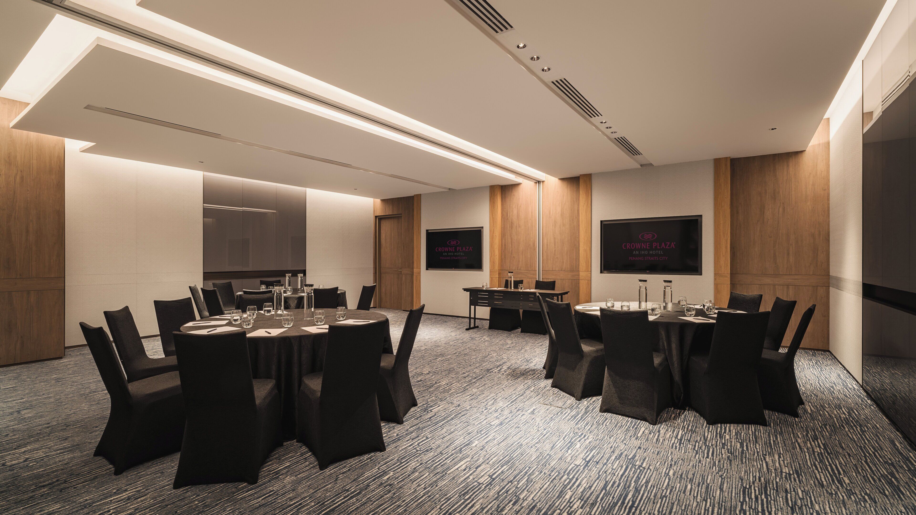 Ein Konferenzraum mit runden Tischen, Stühlen und einem großen Bildschirm, der den Namen des Hotels zeigt.