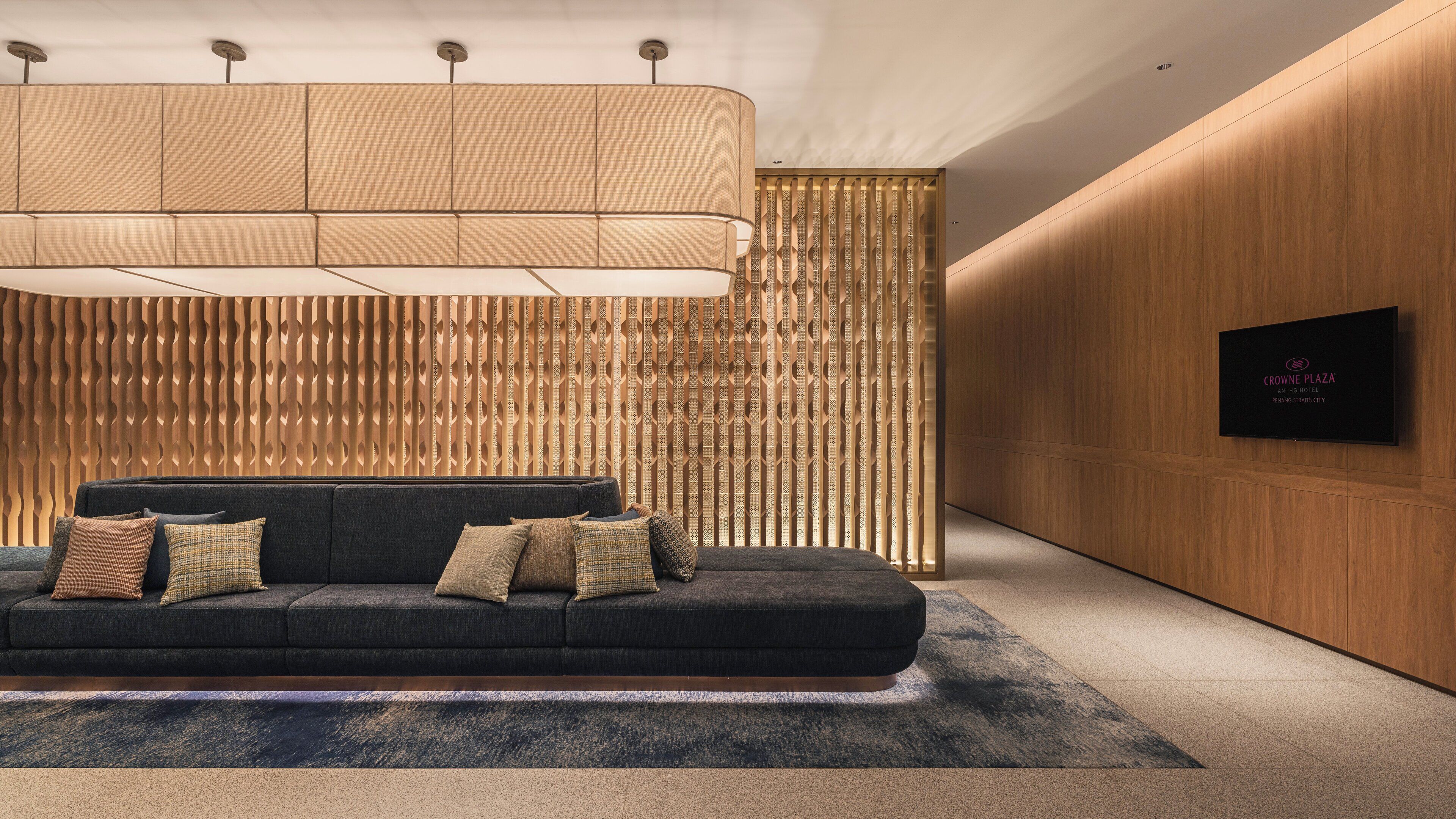 A modern hotel lobby with a dark sofa, patterned cushions, a large wall-mounted TV, and a decorative wooden panel wall.