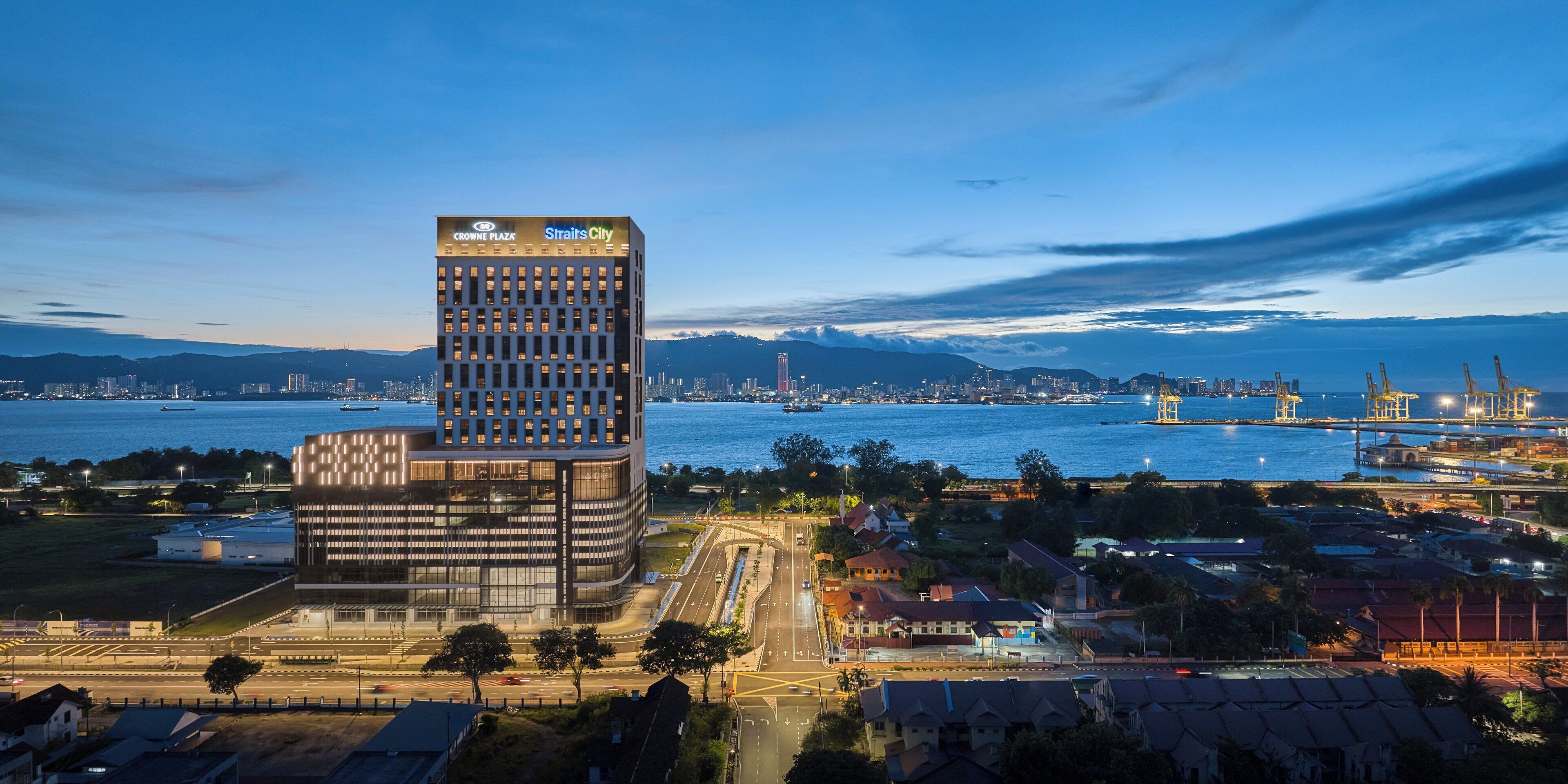 A modern building with a sign reading "Crowne Plaza Straits City" near a waterfront with a view of the city skyline and mountains in the background.