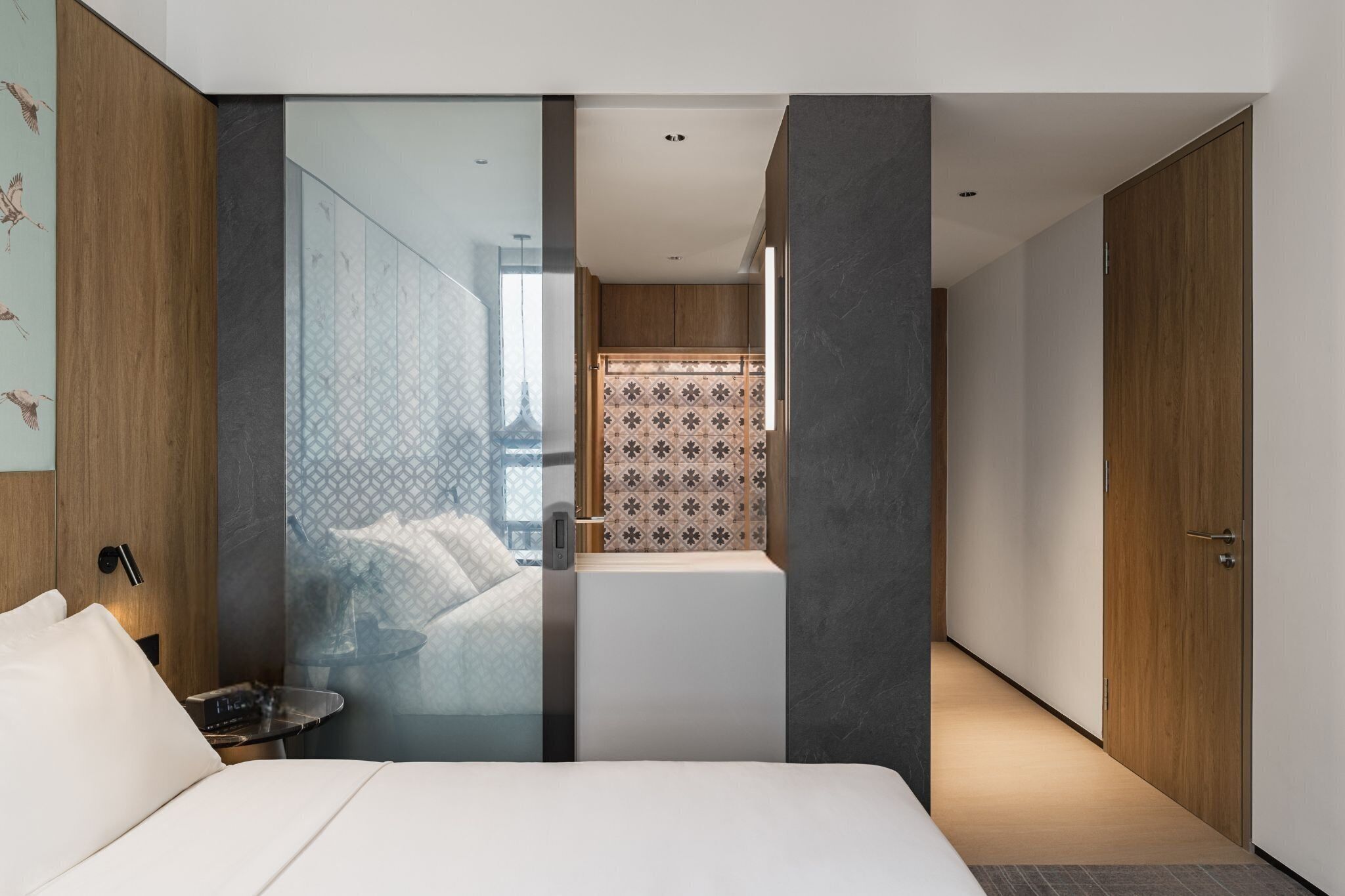 A modern hotel room with a large bed, a bedside table, a bathroom with a shower, and a view of the cityscape.