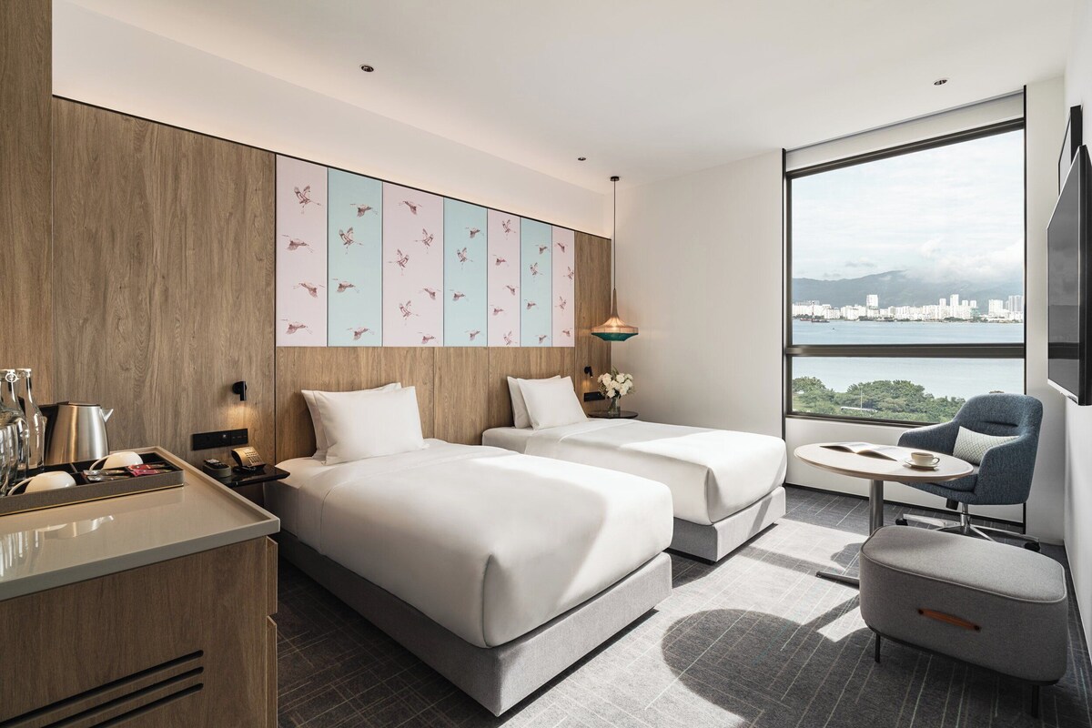 Una habitación de hotel moderna con dos camas, un escritorio pequeño, una silla y un ventanal con vistas a la ciudad.