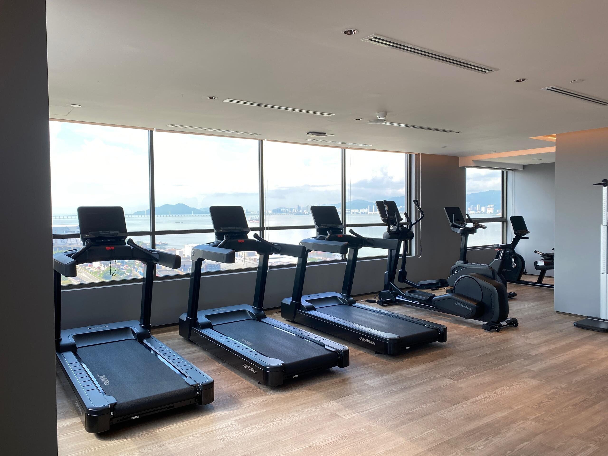 A gym with treadmills aligned against a wall with large windows overlooking a cityscape.