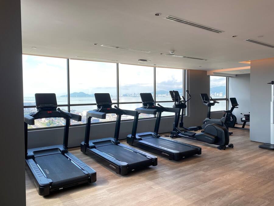 Un gimnasio con cintas de correr alineadas a lo largo de una pared con ventanas amplias que dan a un paisaje urbano.