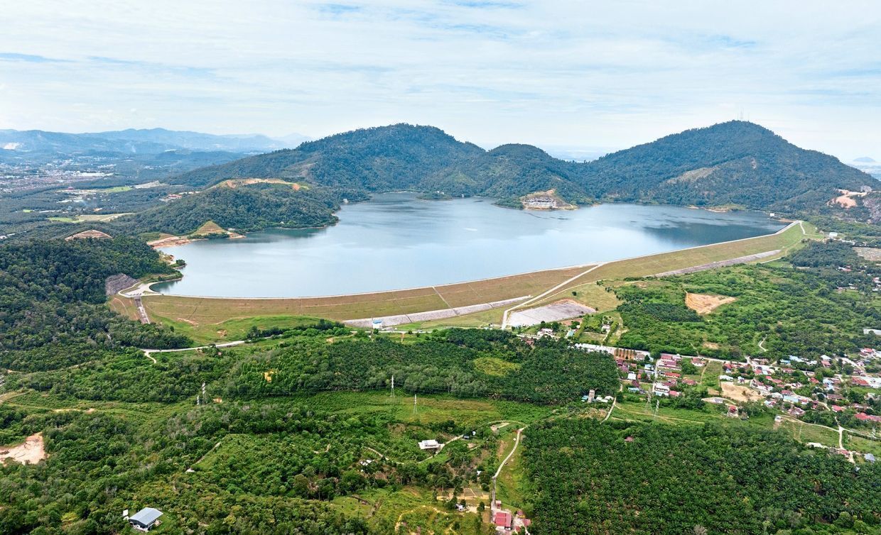 Vista aerea di un lago circondato da colline, con un piccolo paese e una diga in primo piano.