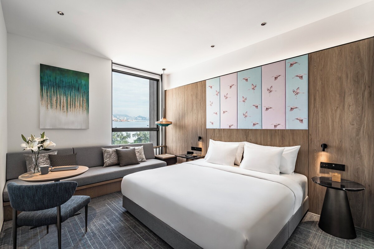 Una habitación de hotel moderna con una cama grande, una zona de estar con un sofá y sillones, una mesita con una lámpara, y un ventanal con vistas a la ciudad.