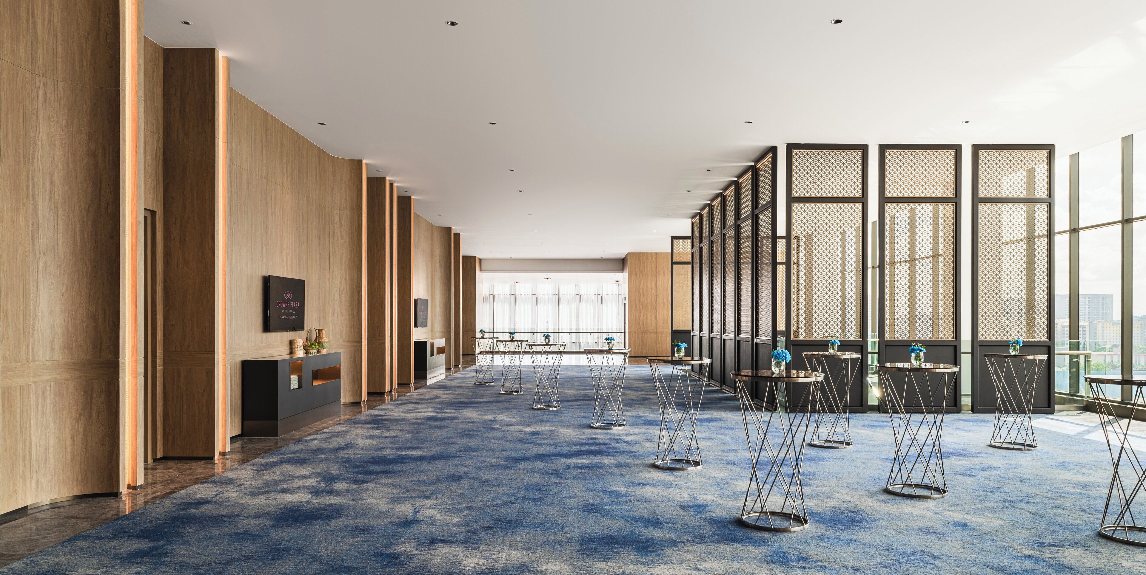 A spacious hotel lobby with a blue carpet, wooden panel walls, and a view of the city through large windows.