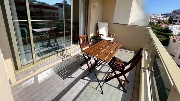 Un balcon avec une table et des chaises en bois, une chaise blanche, et une vue sur les immeubles à travers de grandes portes vitrées.