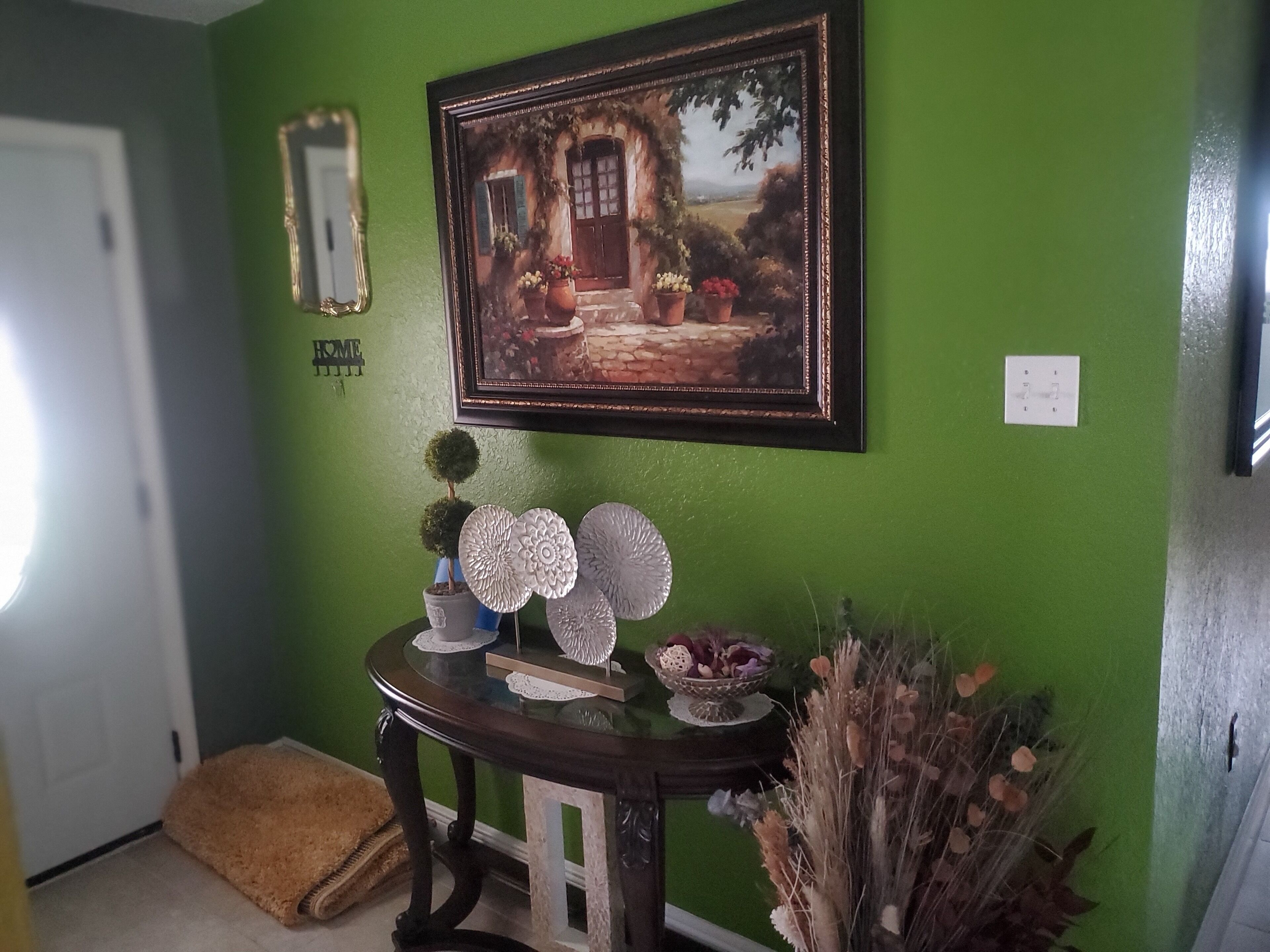 A room with a green wall, a framed painting of a cottage, a small table with decorative items, and a door with a 'HOME' sign.