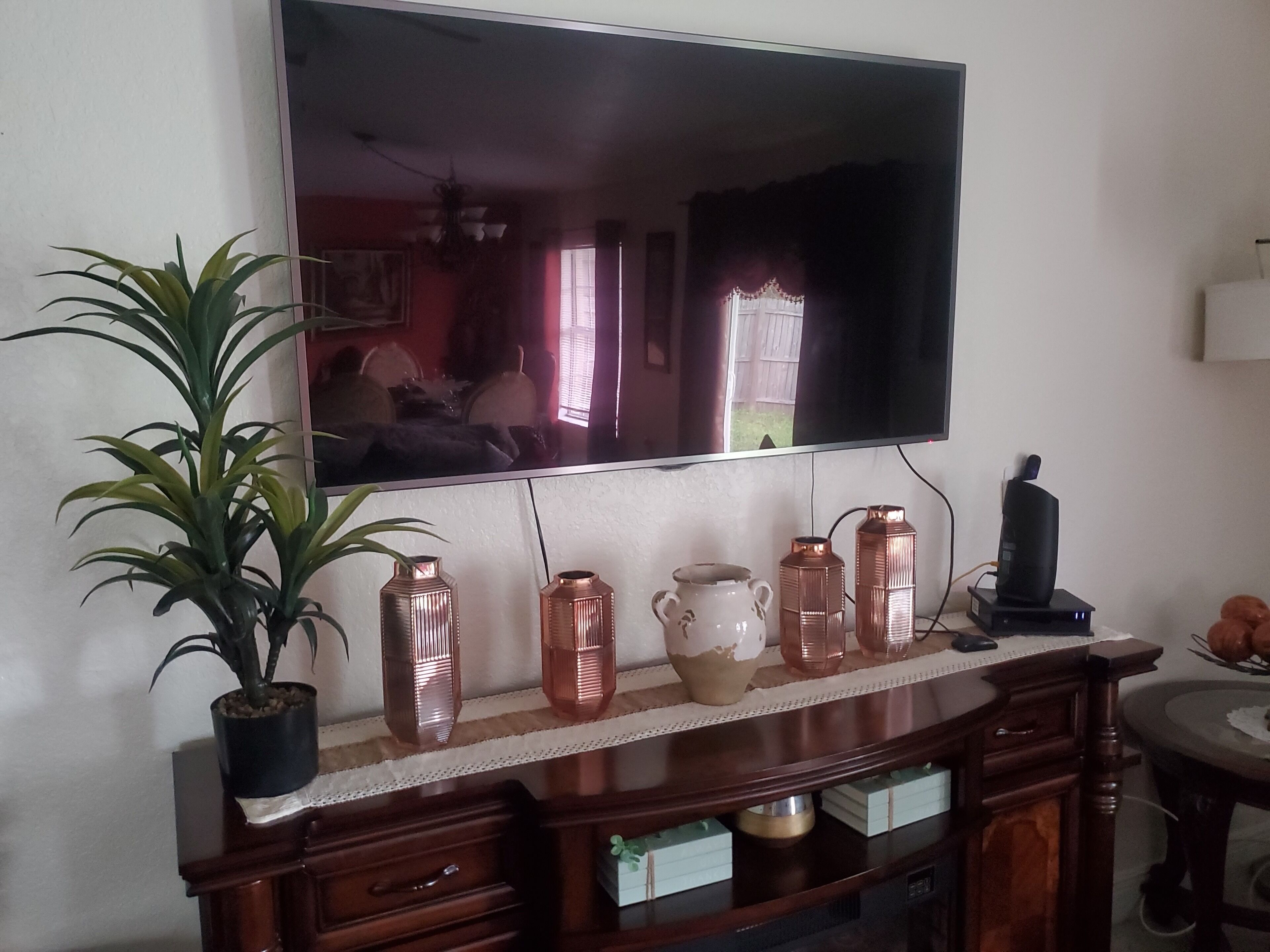 Una sala de estar con una televisión de pantalla plana montada en la pared, una consola de madera con jarrones decorativos, una planta en maceta y un televisor sobre un mueble bajo.