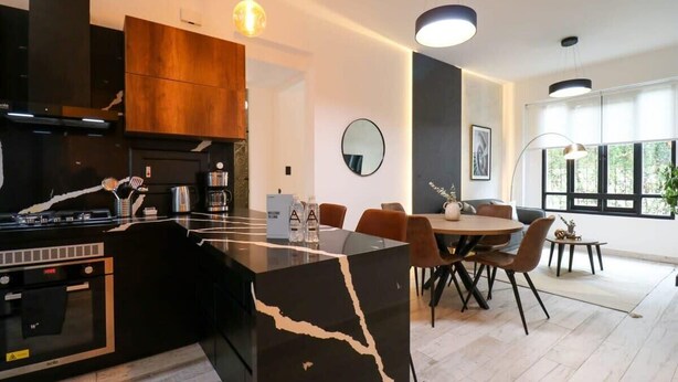 A modern kitchen with a black marble island, wooden cabinets, and a built-in oven. There is a dining area with a round table and chairs, and a large window providing natural light. - w* | Fancy 2BR in Barranco (Barranco)