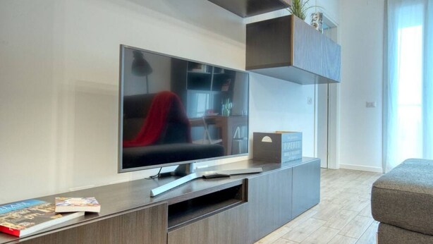 A modern living room with a flat-screen TV mounted on a wooden entertainment unit, a grey sofa, and a small plant on a shelf. - CaseOspitali - Casa Francesca modernissimo trilo (Pioltello)