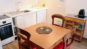 Eine kompakte Küche mit Geschirrspüler, Backofen und Kaffeemaschine. Ein Holztisch mit zwei Stühlen und Untersetzer.
