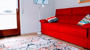 Ein rotes Sofa mit einem gemusterten Zierkissen, eine Stehlampe und ein Teppich in einem Zimmer mit einer Tür und einem gerahmten Kunstwerk an der Wand.