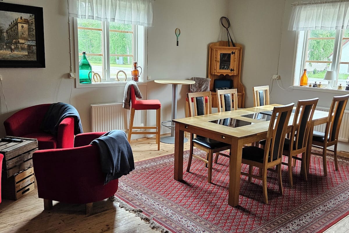 A cozy living room with a red armchair, a wooden dining table with chairs, a radiator, and a large window with curtains.