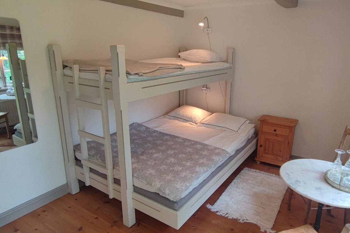 A bunk bed room with a small table and a chair, a window with a view, and a lamp.