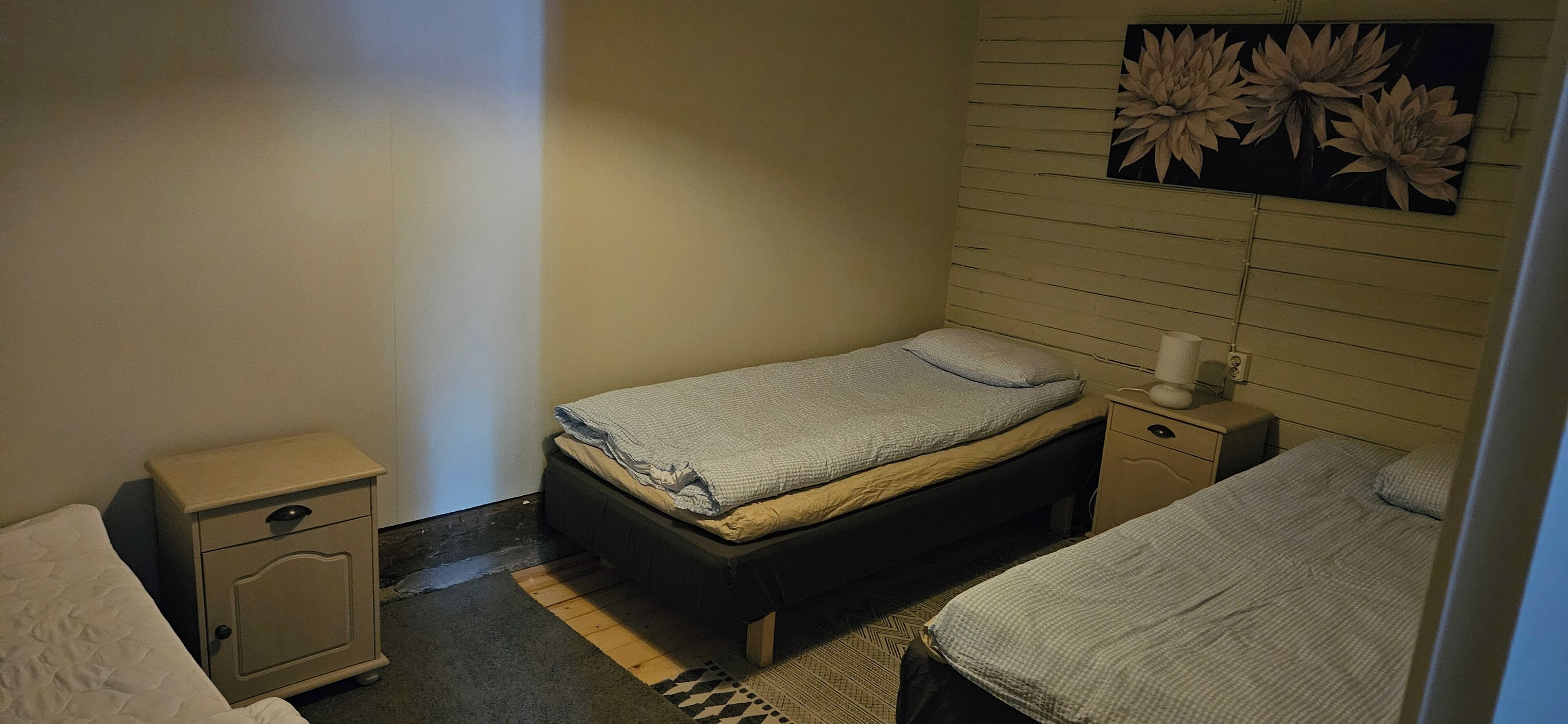 A small, dimly lit room with a single bed, a nightstand, and a wall-mounted flower decoration.