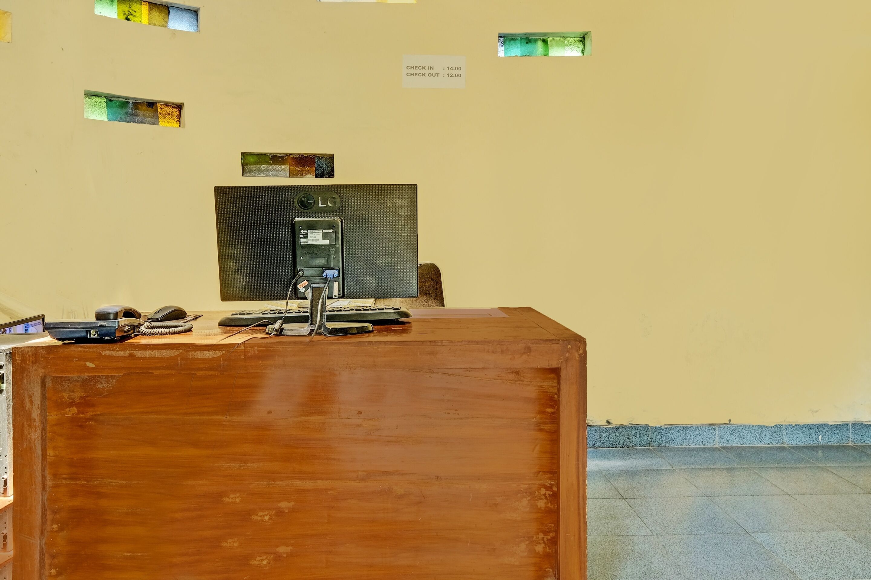 A reception desk with a computer, phone, and keyboard. There are small windows on the wall and a sign indicating check-in and check-out times.