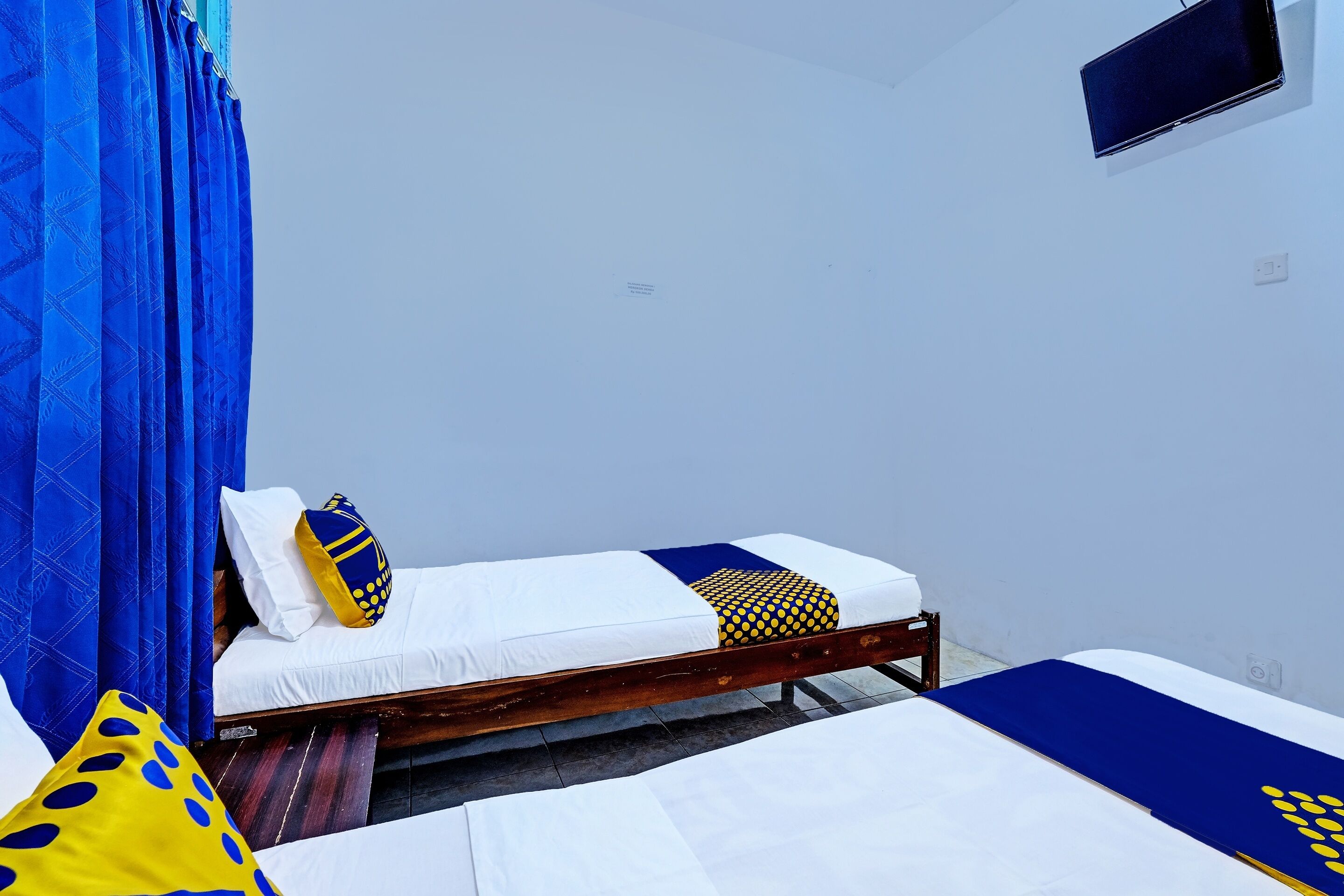 A room with two beds, one with a blue and white bedspread and the other with a white bedspread. There is a TV mounted on the wall above the beds.