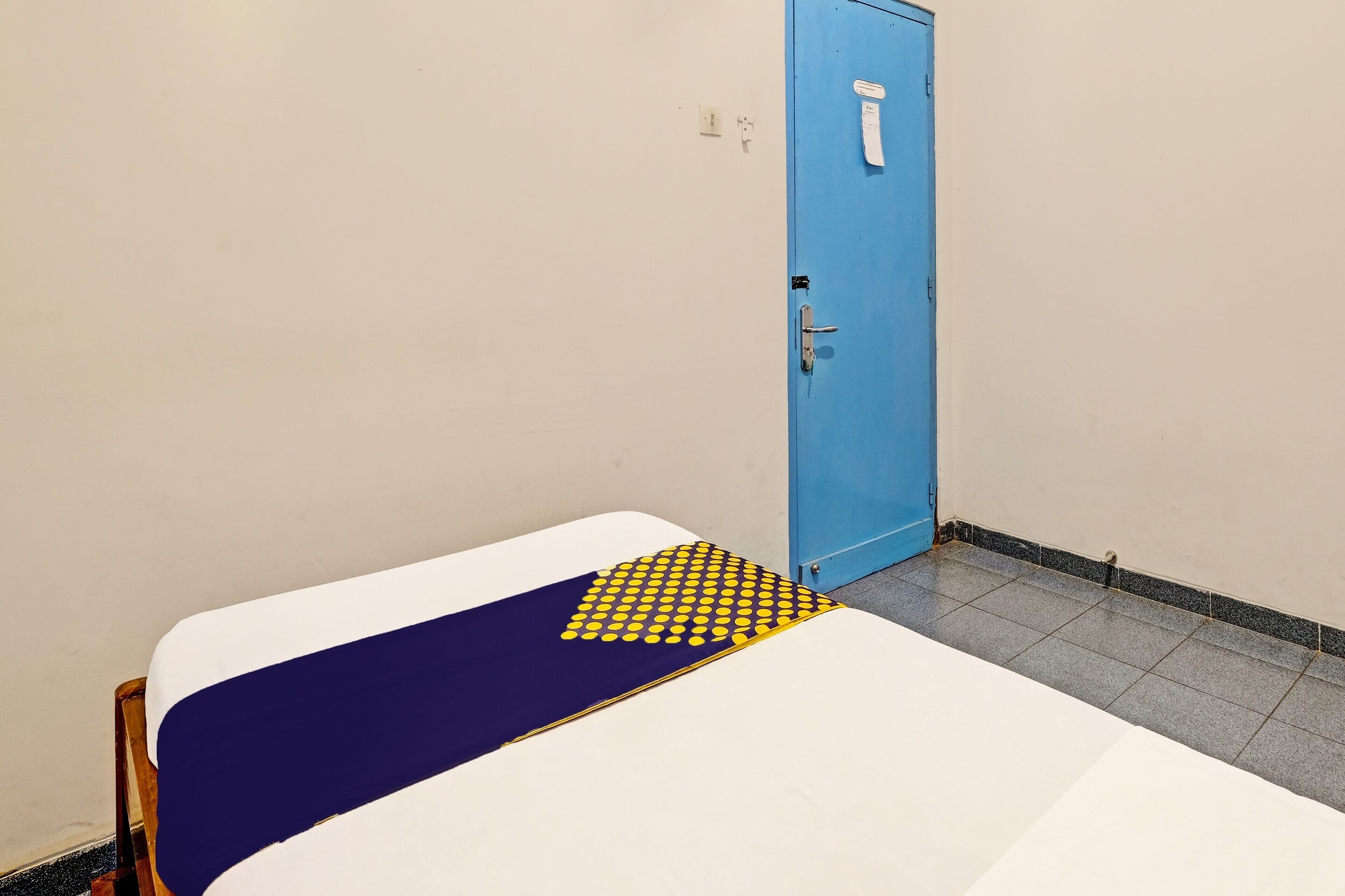 A single bed with a blue and white bedspread, a blue door, and a tiled floor.