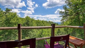 A wooden deck with two chairs and a table, overlooking a forested area with a clear sky.