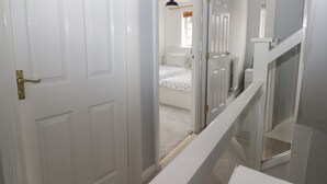 Un pasillo con puertas blancas, una escalera y un dormitorio con cama y ventana.