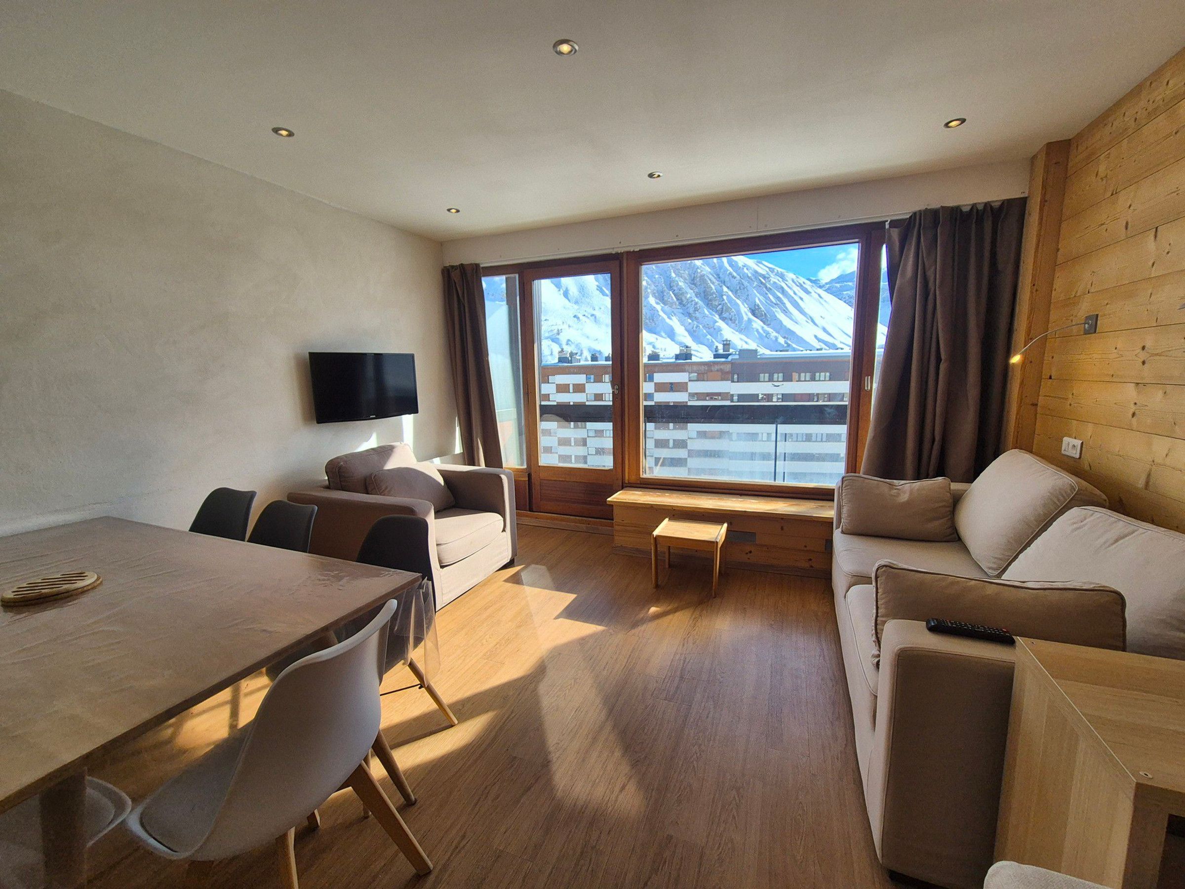 Une chambre d’hôtel moderne avec un plancher en bois, une table à manger avec des chaises, un canapé, un téléviseur fixé au mur et de grandes fenêtres offrant une vue sur les montagnes.