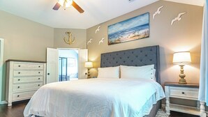 Een slaapkamer met een groot bed, een dressoir, een nachtkastje met een lamp, een plafondventilator en een schilderij van een strand.