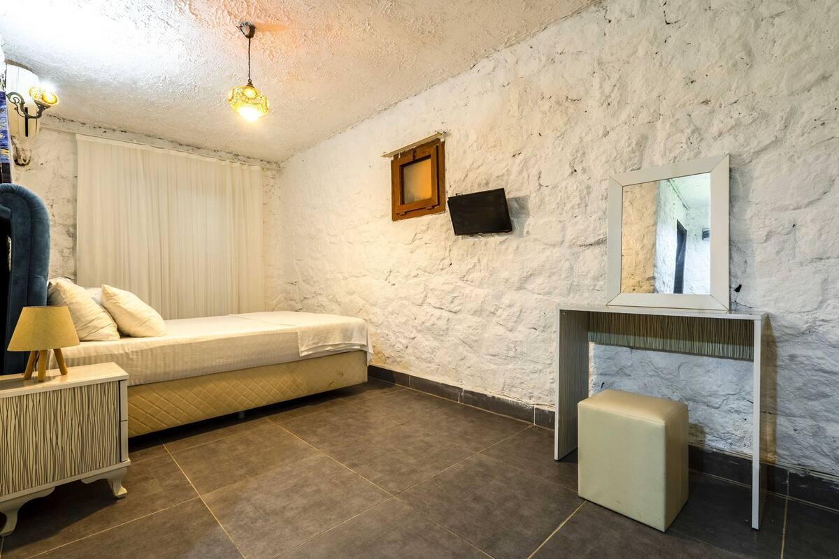 A bedroom with a bed, bedside table, lamp, and a small stool. There is a wall-mounted TV and a mirror above a desk.