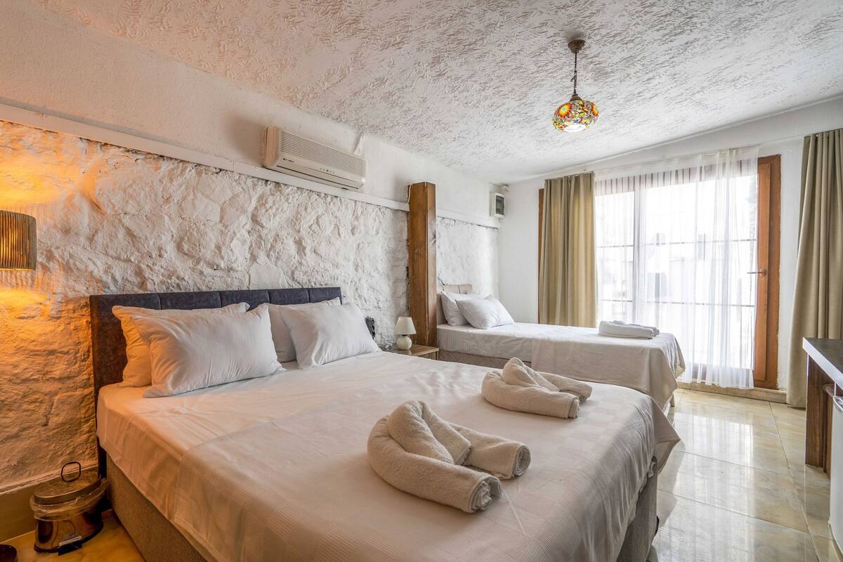 A hotel room with a stone-textured wall, a bed with white linens, two bedside tables, a lamp, and a large window with curtains.