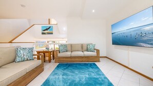 A living room with a sofa, a coffee table, and a blue rug. There is a painting on the wall and a large window with a view of the sea.
