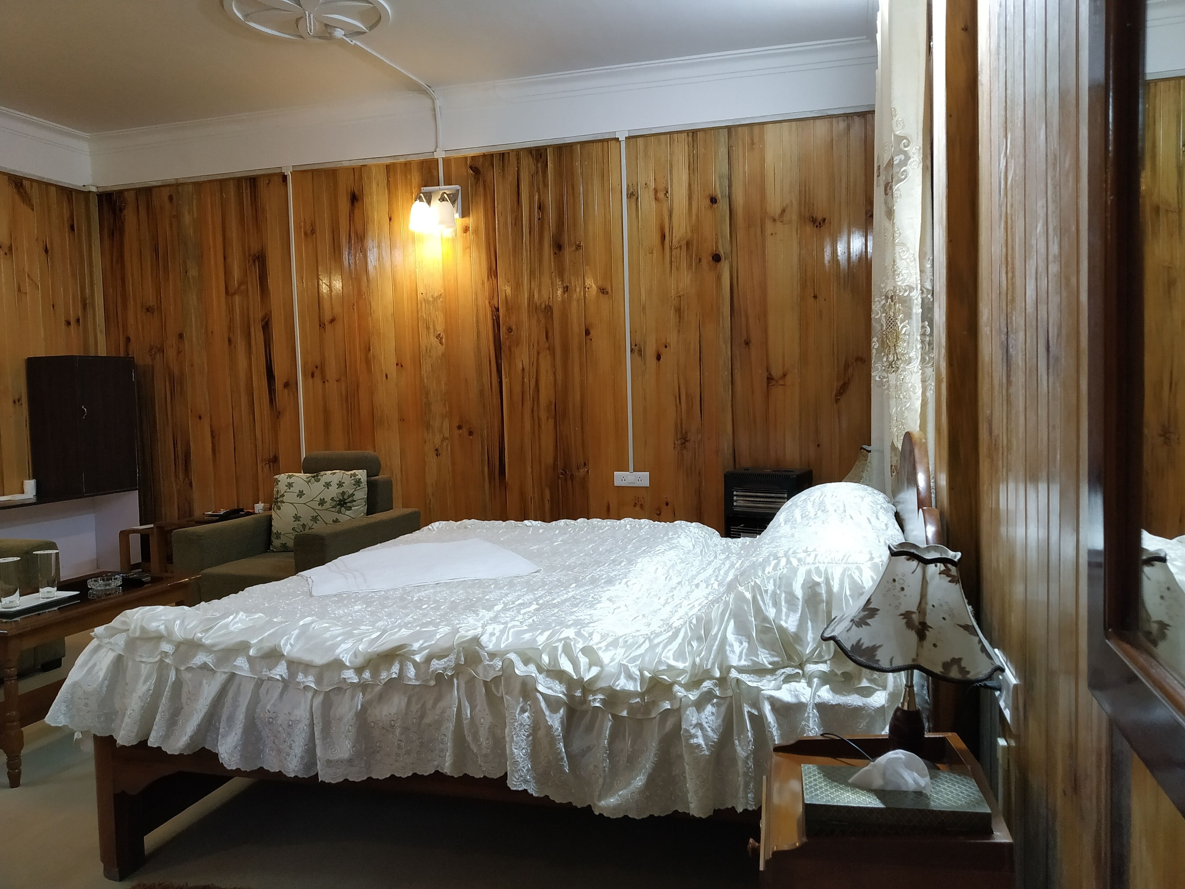 A room with a wooden paneled wall, a bed with a white bedspread, a small table, a chair, and a lamp.