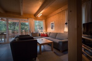 Relax in this inviting living room with a sofa set, coffee table, and TV on a wooden stand, offering a view of the outdoors through large windows.