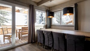 A modern dining area with a long table and chairs, large windows, and a hanging light fixture.