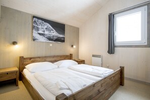 Una camera da letto con un letto di legno, un comodino, una lampada a parete, una finestra con tende e un dipinto con un paesaggio montano.