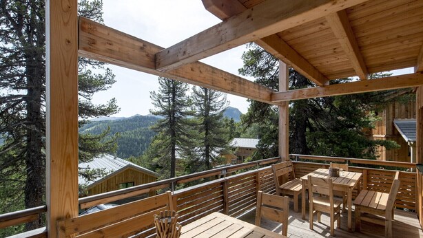 A wooden deck with tables and chairs, overlooking a forested area with mountains in the background.