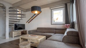 A modern living room with a sectional sofa, a wooden coffee table, a TV on a wooden stand, a hanging lamp, and a staircase with a view to the outside.