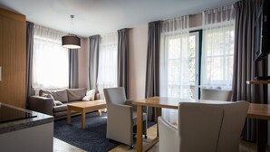 Ein modernes Wohnzimmer mit einer Couch, Sesseln, einem Couchtisch und einem Esstisch. Ein großes Fenster mit Vorhängen, eine Deckenleuchte und ein Fernseher auf einem Lowboard.