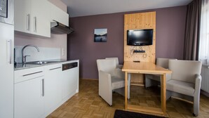 Une cuisine compacte équipée d’un évier, d’un lave-vaisselle et d’un téléviseur à écran plat fixé sur un panneau en bois. Le coin salon comprend deux chaises et une petite table.