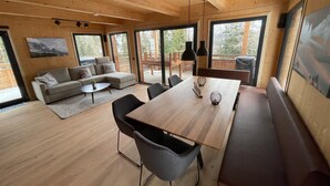 Ein modernes Interieur mit Holzbalkendecke, großen Fenstern, einer Couch, einem Esstisch und einer Bank.