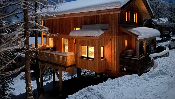 Eine Holzhütte mit Balkon, großen Fenstern und einem mit Schnee bedeckten Dach.