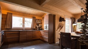 A rustic cabin interior with wooden walls and ceiling, a kitchen with a built-in oven, a dining area with a table and chairs, and a large window with curtains. - Chalet #16 with IR-sauna and indoor whirlpool (Turrach)