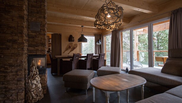 A cozy cabin interior with a stone fireplace, wooden ceiling beams, and a large window overlooking a forest. - Chalet #16 with IR-sauna and indoor whirlpool (Turrach)