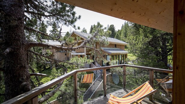 A wooden deck with a hammock chair, overlooking a forested area with multiple buildings. - Chalet # 20 with IR-sauna and whirlpool bath (Turrach)
